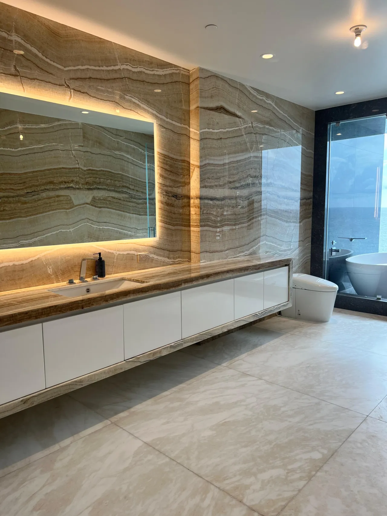 Bathroom with a book-matched veined-stone accent wall, a backlit mirror, and a floating stone-slab vanity with white wall cabinets