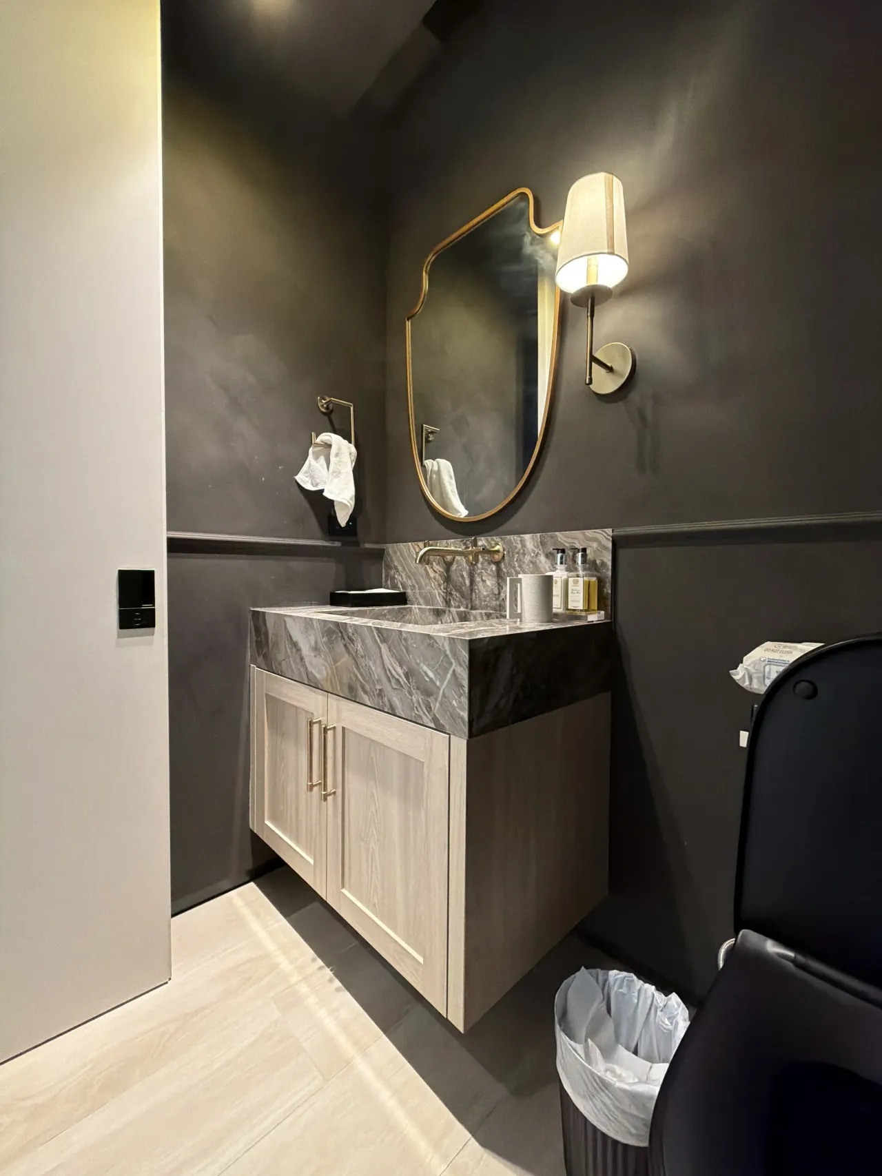 Dark charcoal walls with a shaped gold-tone framed mirror, a shaded sconce, and a gray-veined stone vanity top on a wood-grain melamine cabinet