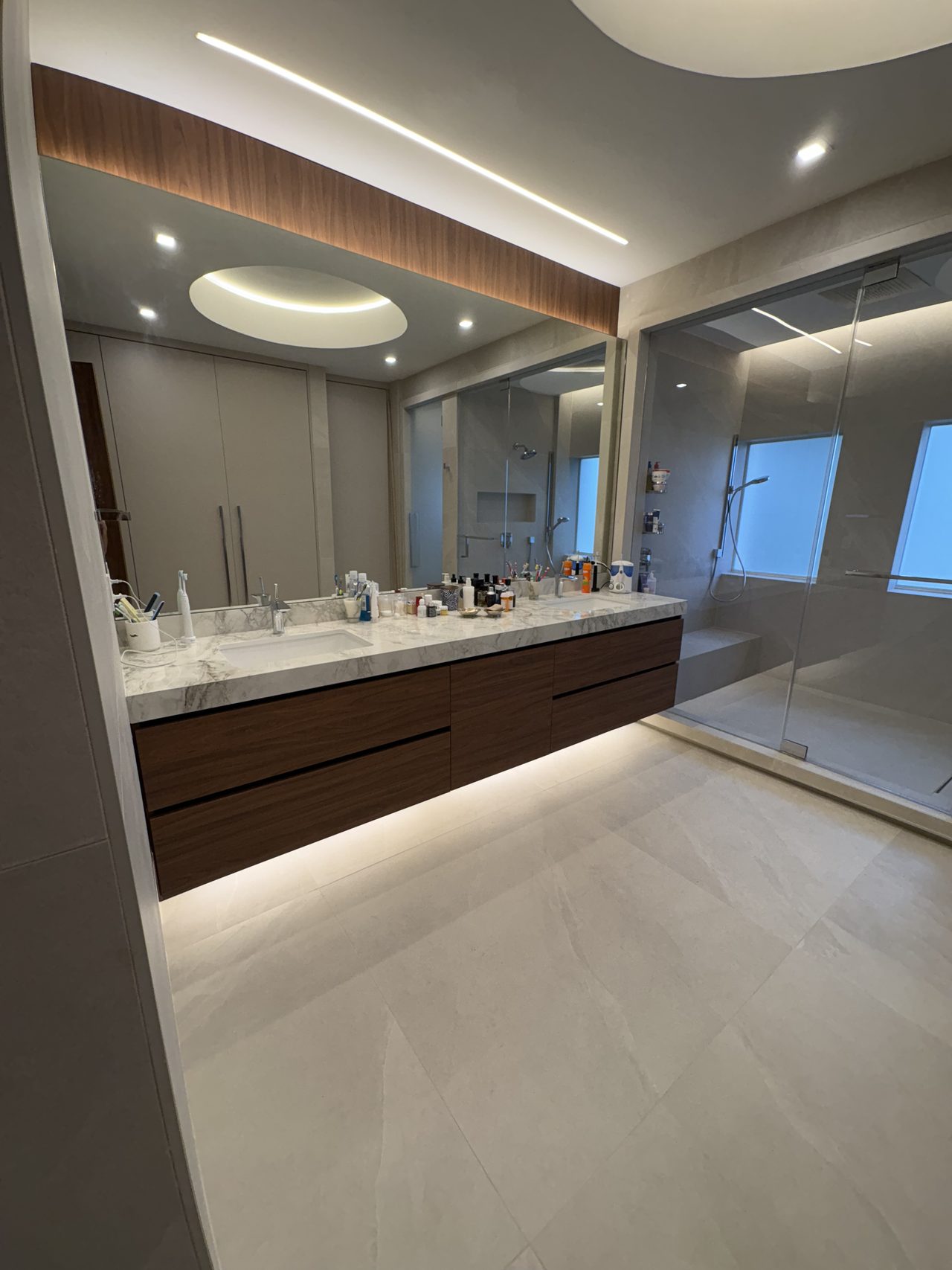 Dark wood-grain melamine floating double vanity with a white stone countertop, a large frameless mirror with a wood-clad soffit, and a glass shower beyond