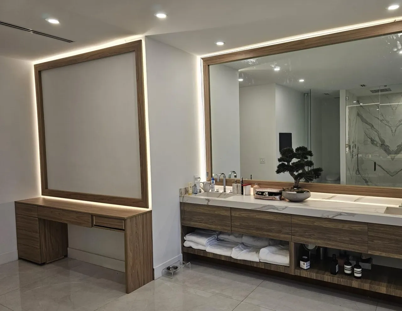 Dark wood-grain vanity run with a veined stone top and an open shelf of folded towels, under a large backlit wood-framed mirror, next to a wood desk