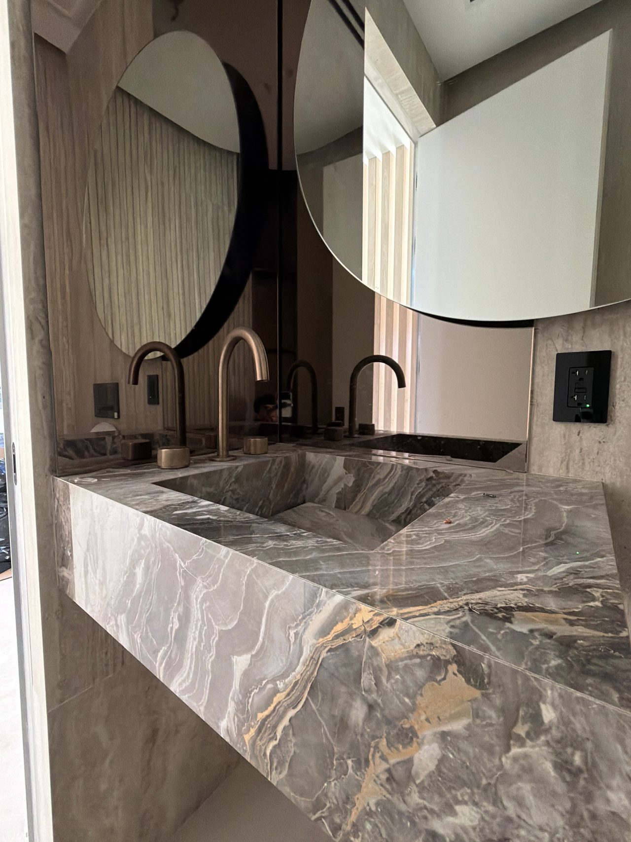 Deck-mounted metallic gooseneck faucet on a thick gray stone vanity with amber veining, under a round mirror and a mirrored backsplash panel