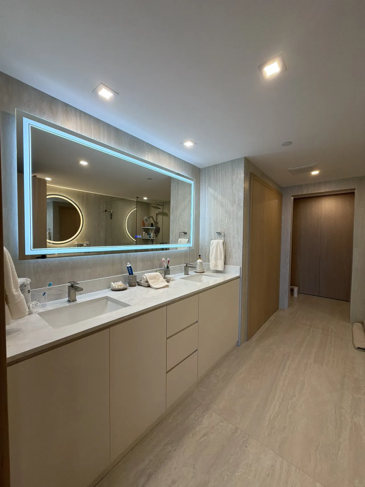 Double-sink vanity with beige cabinetry and a white stone counter under a large rectangular mirror with a glowing blue LED border