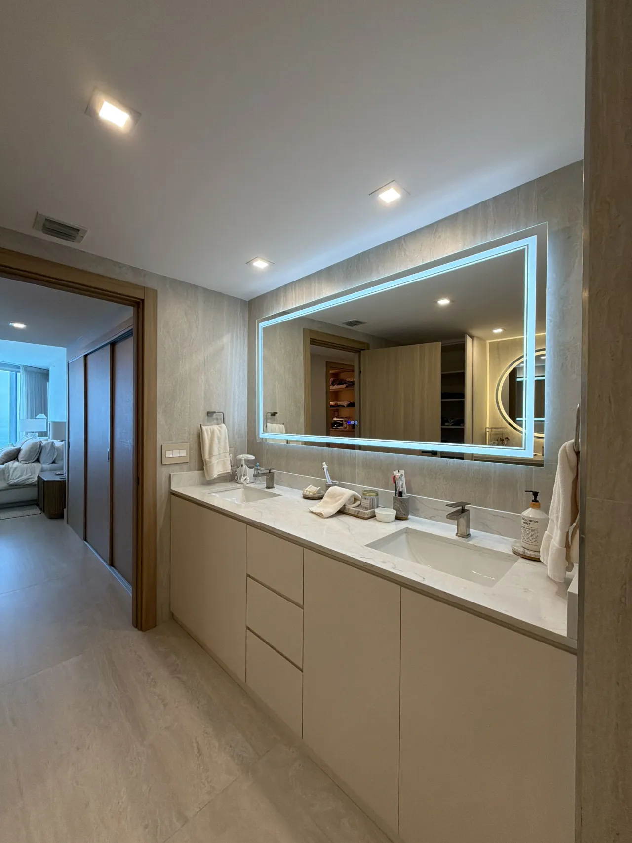Long double-sink bathroom vanity with beige flat-panel cabinets, a veined-stone countertop, and a mirror with a glowing blue-white LED border