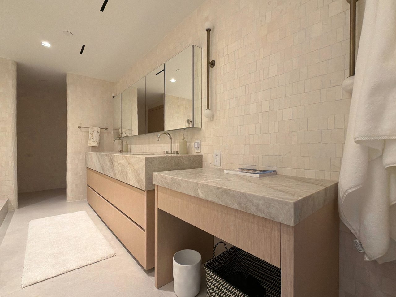 Double vanity with custom-stained wood veneer drawer fronts under a thick veined stone counter with integrated sinks, mirrored cabinets and a seated return
