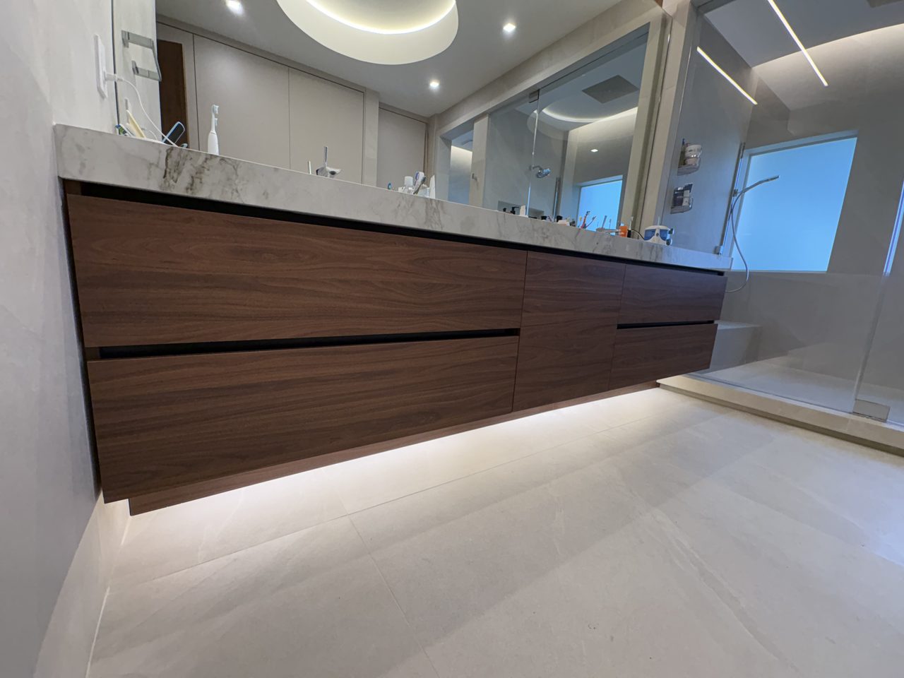 Floating dark wood-grain melamine vanity with two rows of flat drawer fronts, a white veined stone top, and LED glow beneath the cabinet