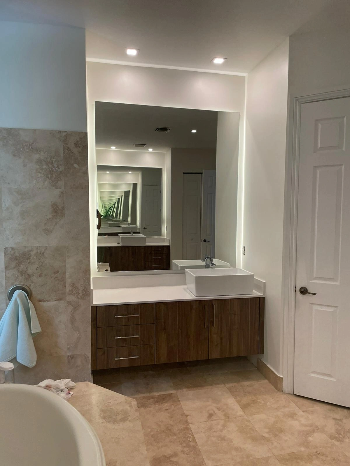 Floating dark wood vanity with a white rectangular vessel sink atop a light stone counter, under a tall backlit mirror, with a soaking tub nearby