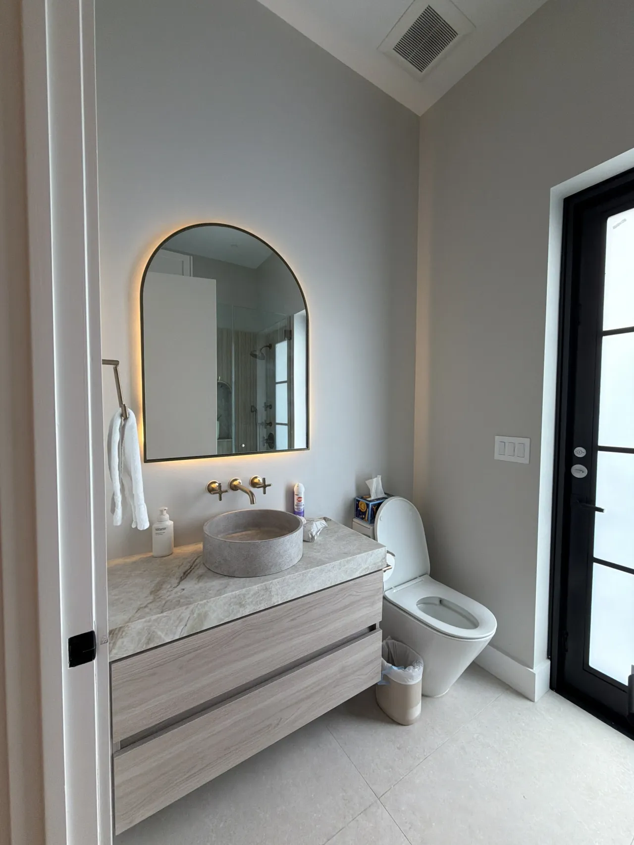 Floating vanity in a light wood-grain melamine finish with a round stone vessel sink and a wall-mounted gold-tone faucet, under an arched backlit mirror