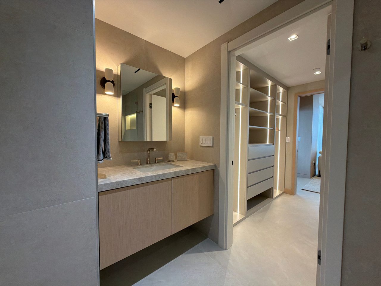 Floating custom-stained wood veneer vanity with a veined stone top and integrated sink, mirrored cabinet between sconces, LED-lit closet shelving beyond