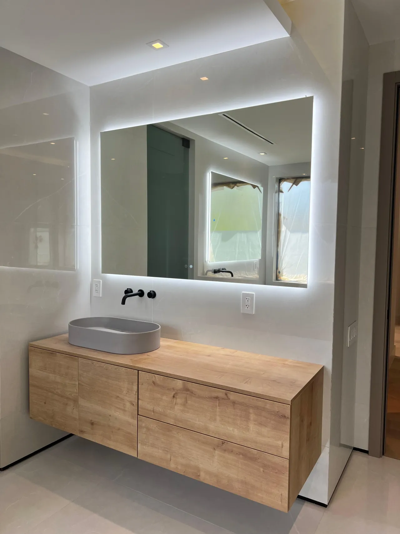 Floating wood-grain vanity with a gray oval vessel sink, a black wall-mount faucet, and a large backlit mirror on a pale stone-look wall