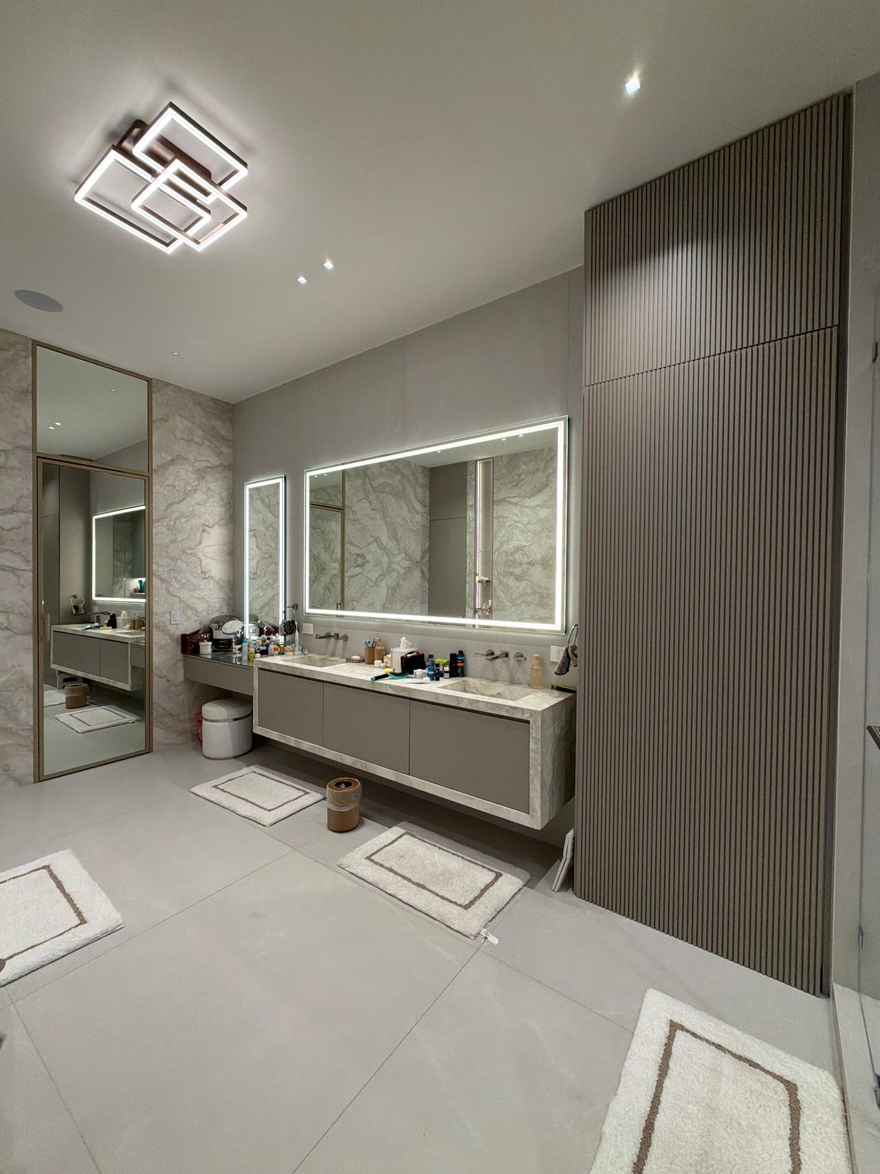 Bathroom with a floor-to-ceiling fluted wood-look wall panel, a taupe floating vanity with stone-look top, backlit LED mirrors, and bath mats
