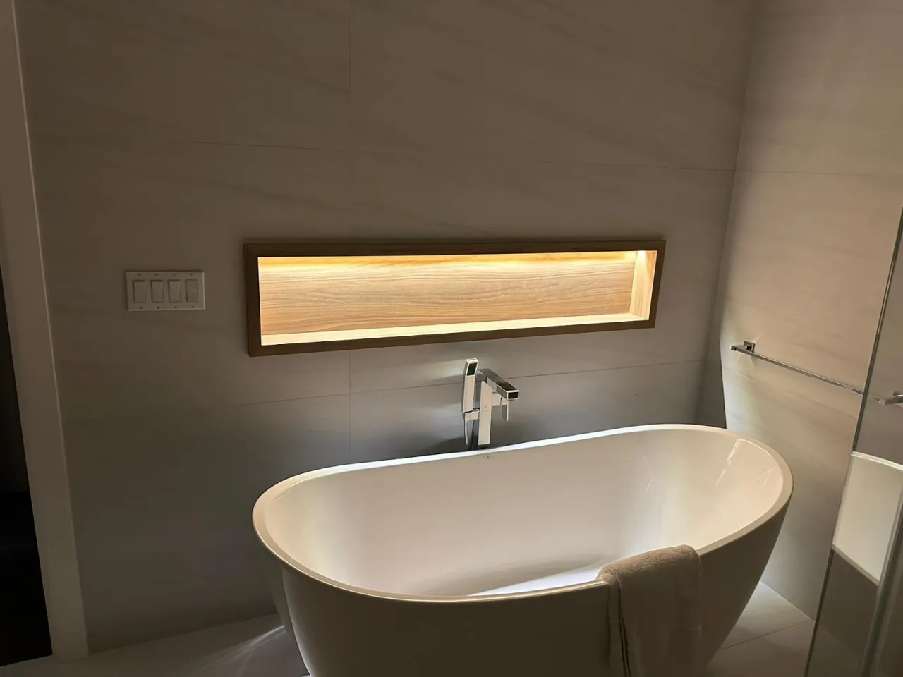 Freestanding oval soaking tub with a tall chrome filler, beneath a long rectangular wood-lined wall niche with warm integrated lighting
