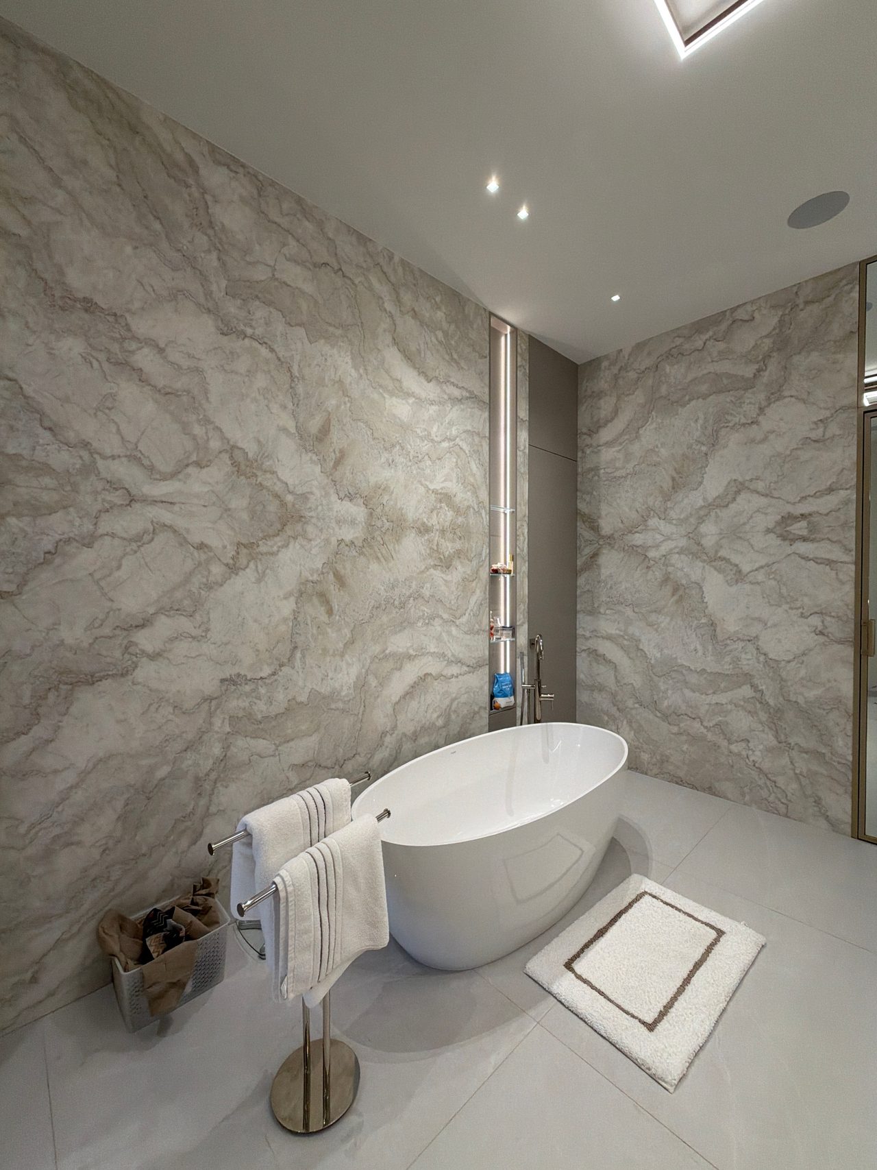 Freestanding oval tub against veined stone slab walls, with a lit recessed niche, a standing towel bar with white towels, and a bath mat
