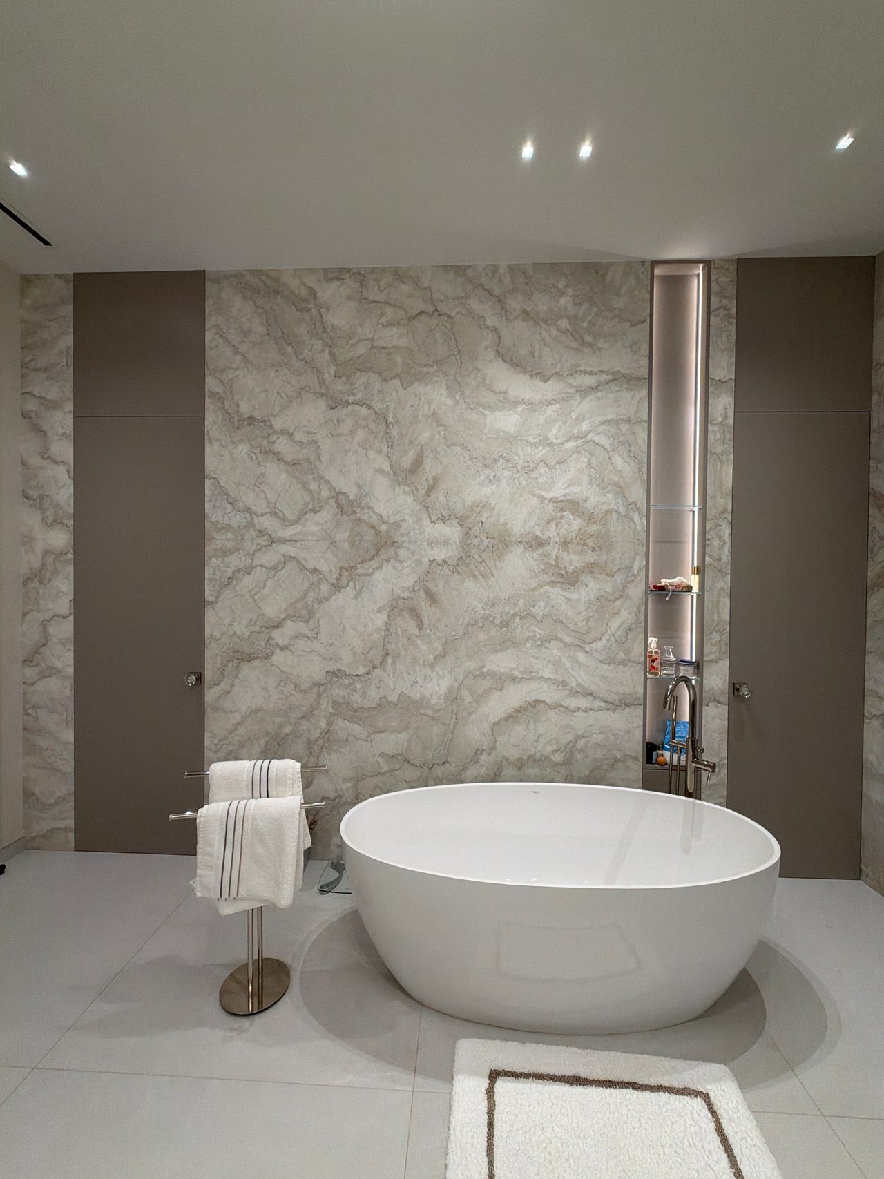 Freestanding oval tub before a veined stone slab wall flanked by taupe flat-panel cabinetry, with a lit niche shelf and a standing towel bar