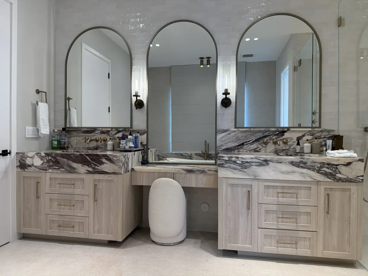 Light wood-grain melamine double vanity with triple arched mirrors, dark-veined stone countertops, and a glass shower visible at right