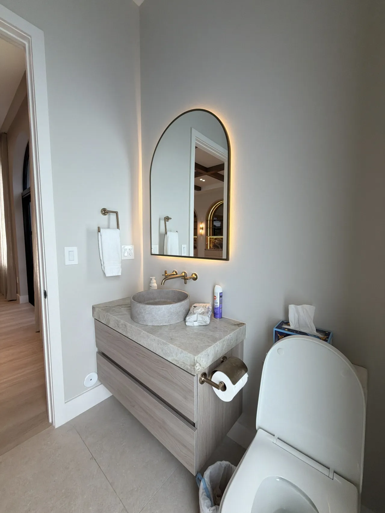 Small powder room with an arched backlit mirror, a stone vessel sink on a wood-grain melamine floating vanity, gold-tone fixtures, next to a toilet
