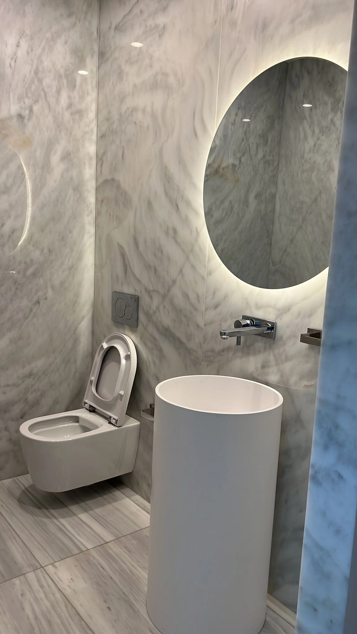 Powder room with a cylindrical freestanding pedestal sink and a wall-hung toilet against gray-veined stone walls beneath a backlit round mirror
