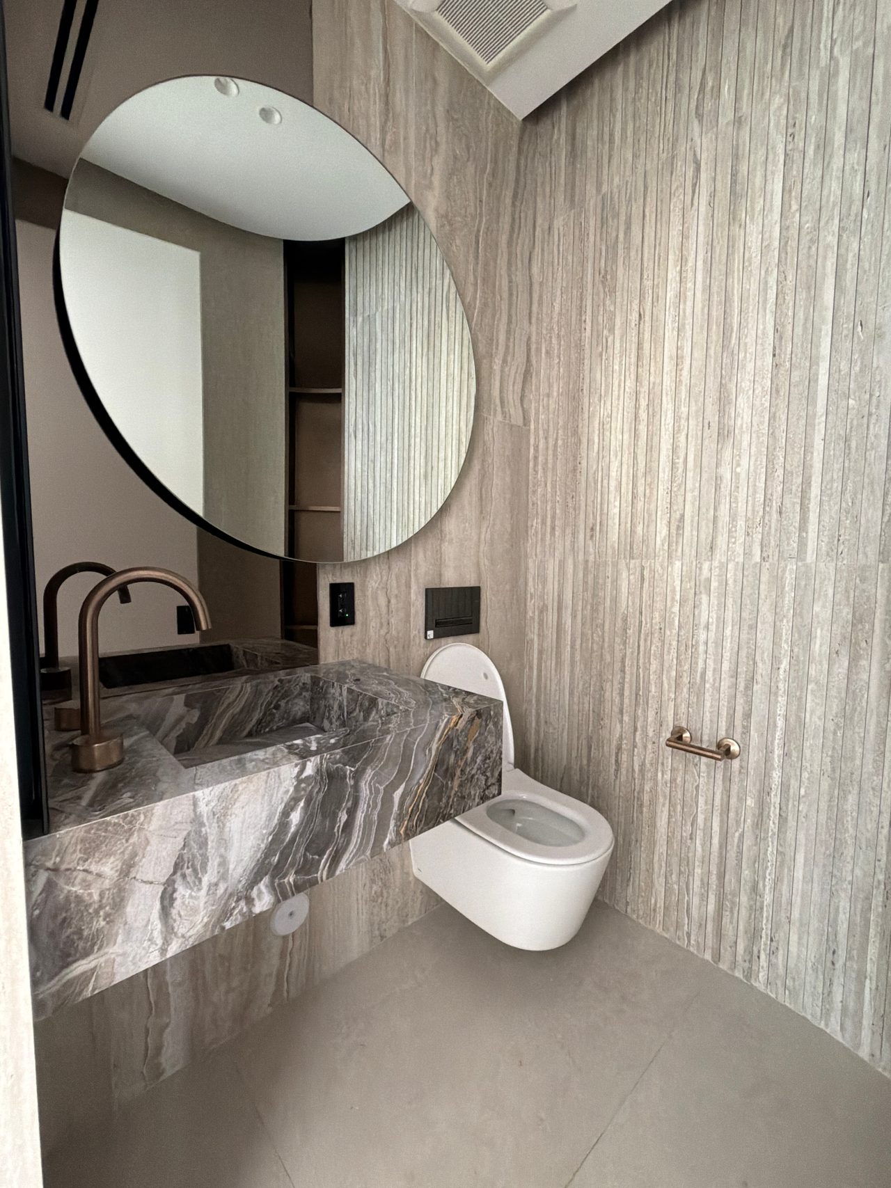 Bathroom with a large round black-framed mirror, vertical striated stone-look wall tile, a floating veined stone vanity, and a wall-hung toilet