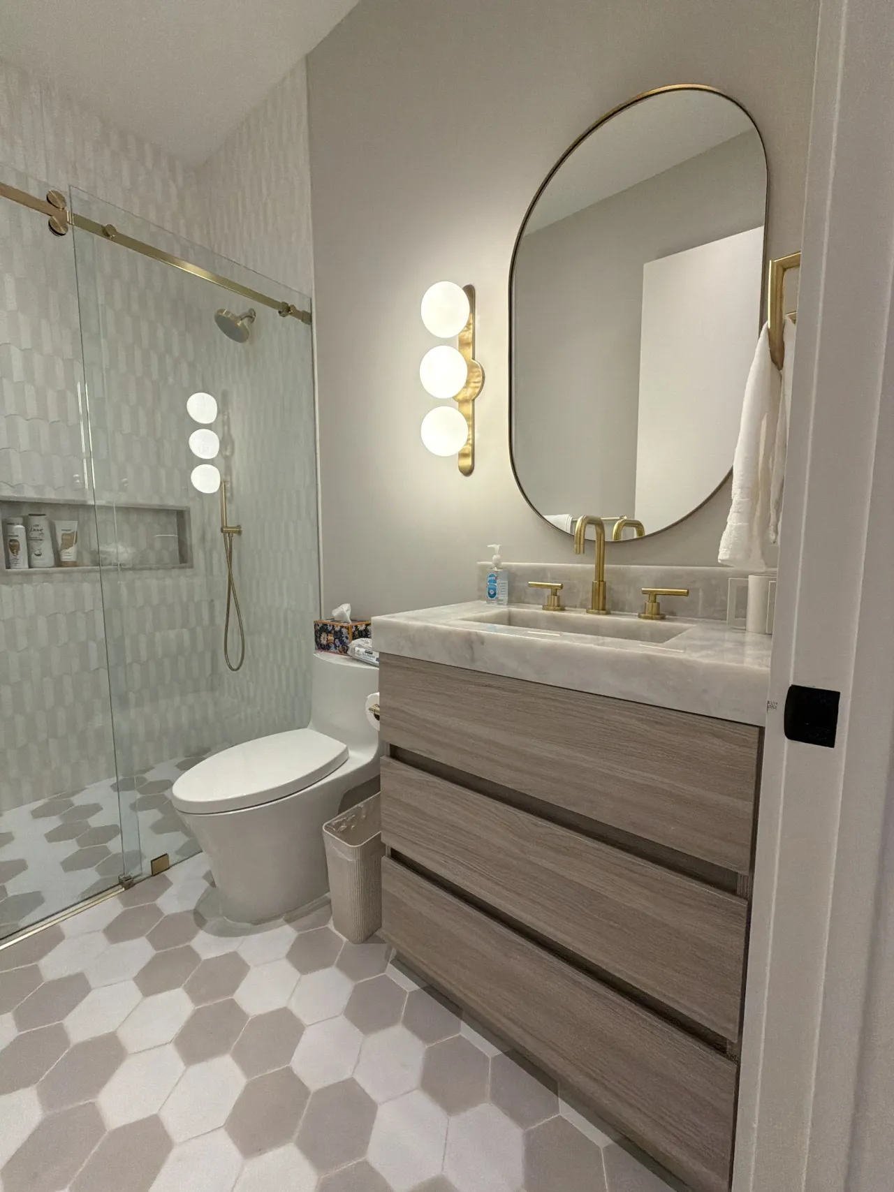 Taupe bathroom with a glass shower and toilet alongside a wood-grain melamine vanity with a globe sconce