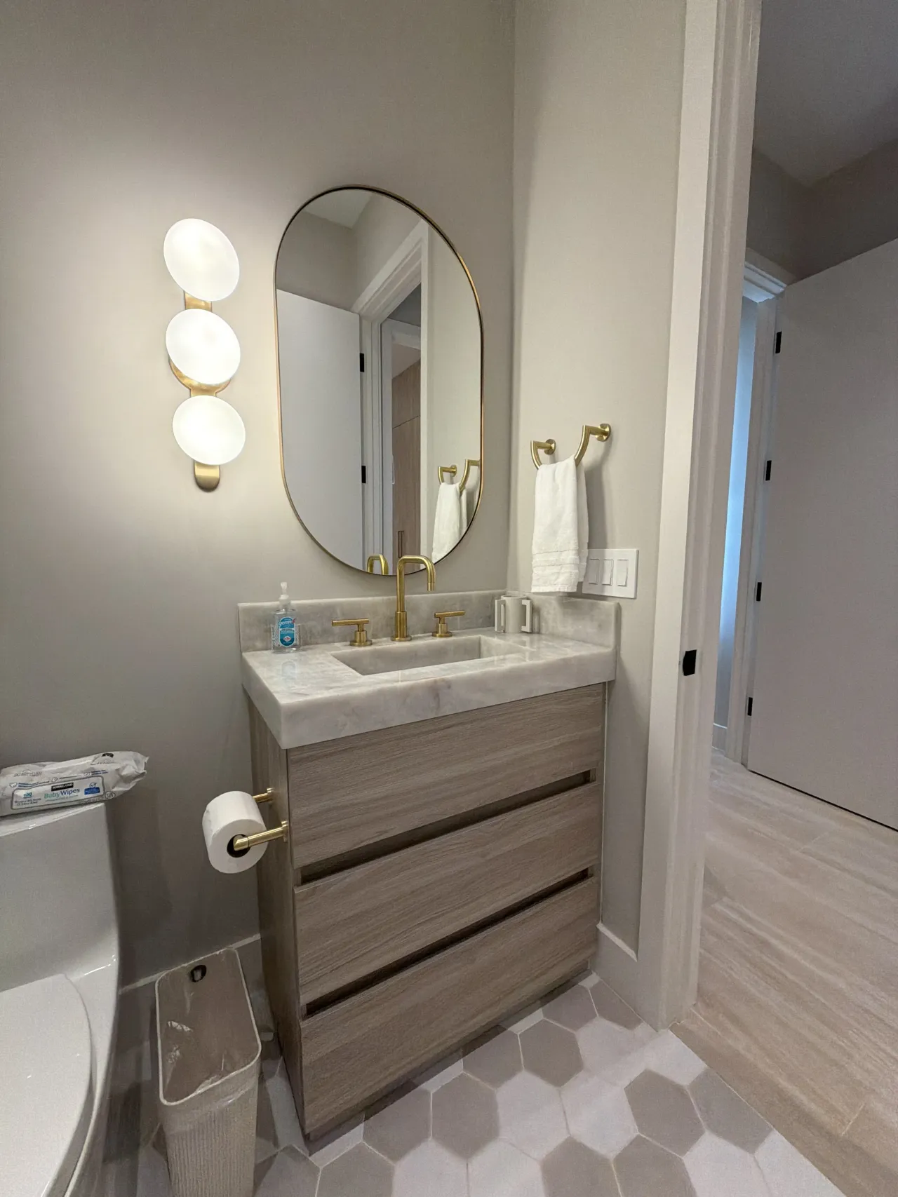 Taupe bathroom with an oval gold-tone framed mirror, a three-globe sconce, and a gray wood-grain melamine vanity with a veined stone countertop