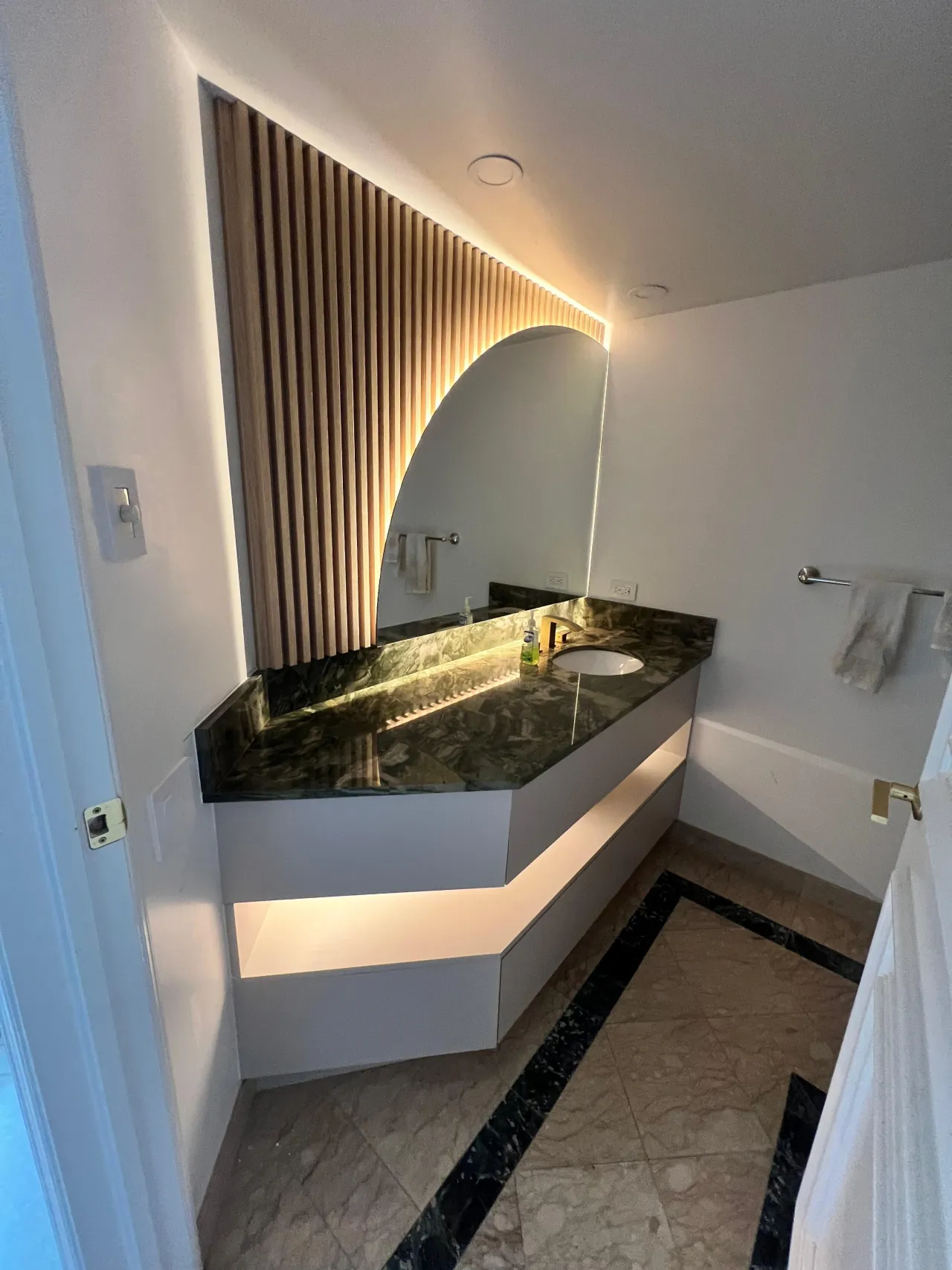 Vanity with a dark green/black stone countertop, a white cabinet base, and an arched backlit mirror set against a vertical wood-slat accent wall