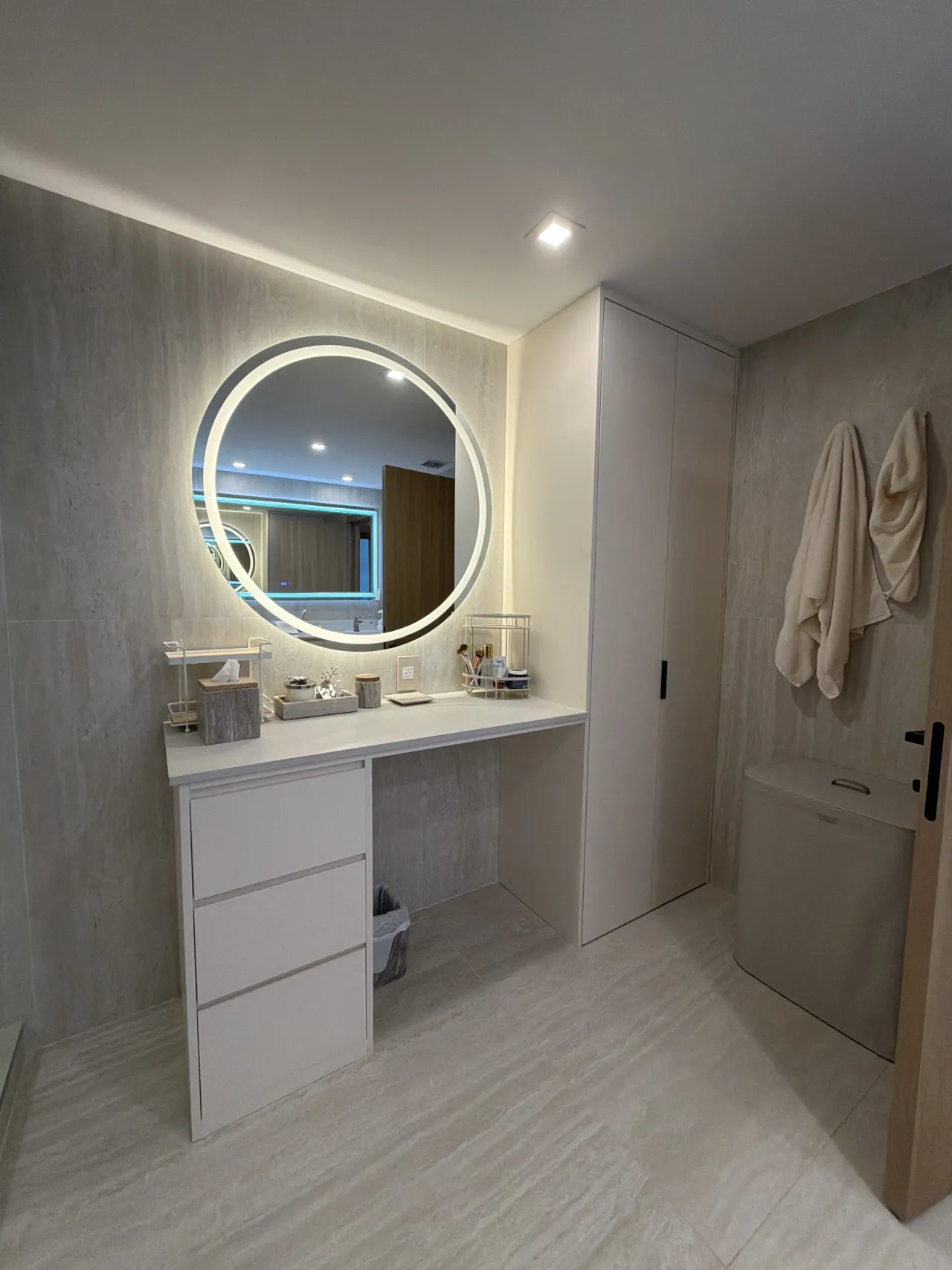 Vanity nook with a round LED-backlit mirror over a white countertop with a knee-space desk area, adjacent to a tall light beige wardrobe/linen cabinet