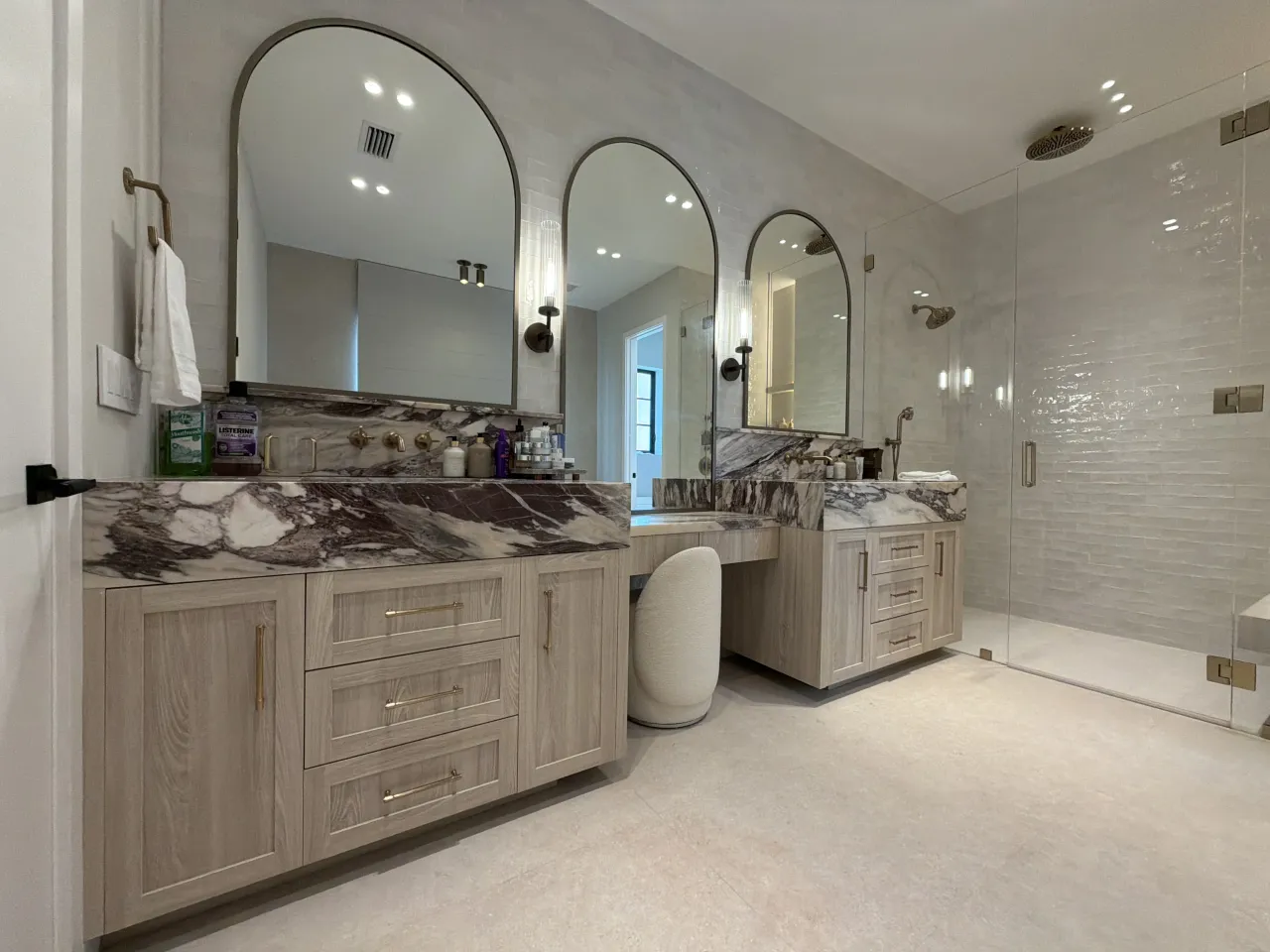 Vanity suite with a glass-enclosed shower with tiled walls and a rainfall showerhead alongside arched-mirror vanities