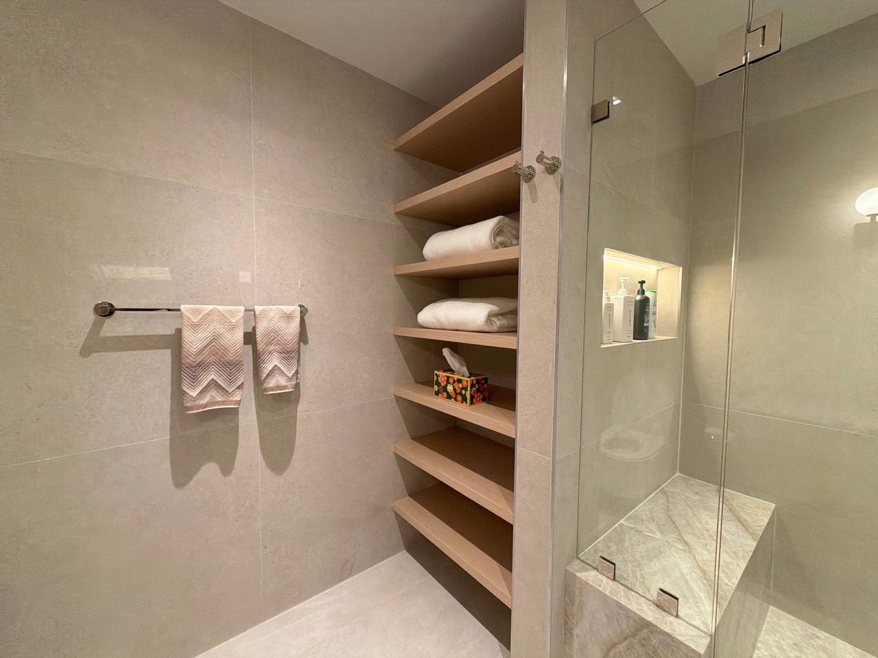 Open custom-stained wood veneer towel shelving built beside a glass shower with an LED-lit recessed niche and a veined stone bench
