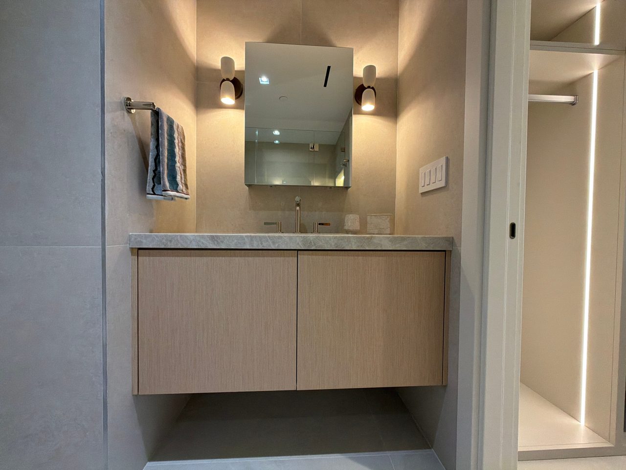 Custom-stained wood veneer two-door floating vanity under a veined stone top, mirrored cabinet flanked by sconces, LED-lit closet shelving at right