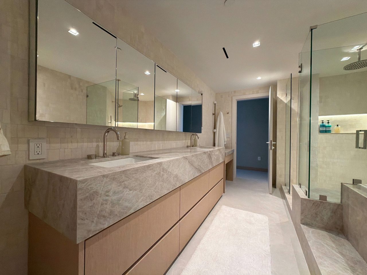 Long custom-stained wood veneer vanity run under a veined stone counter, full-width mirrored medicine cabinets, and a glass shower with a veined stone curb