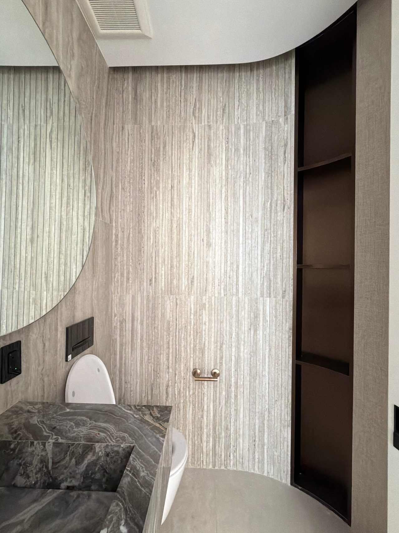 Vertical striated stone-look wall tile with a dark veined stone vanity edge, a wall-hung toilet, and a recessed dark open shelf niche
