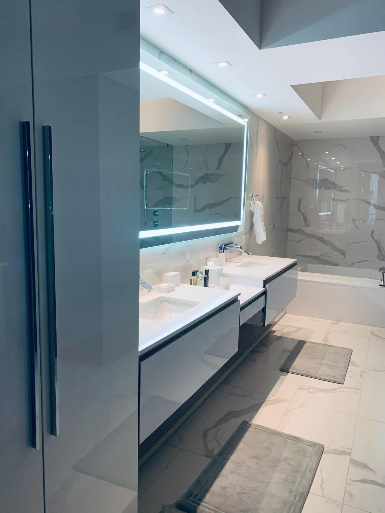White floating double vanity with an integrated sink and stone countertop beneath a large backlit rectangular mirror and gray-veined stone tiled walls