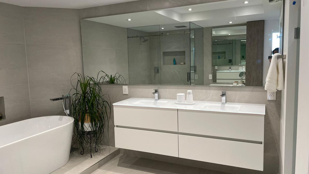 White floating double vanity with white slab tops, gray textured tile walls, an oval freestanding tub, a glass shower, and a potted plant