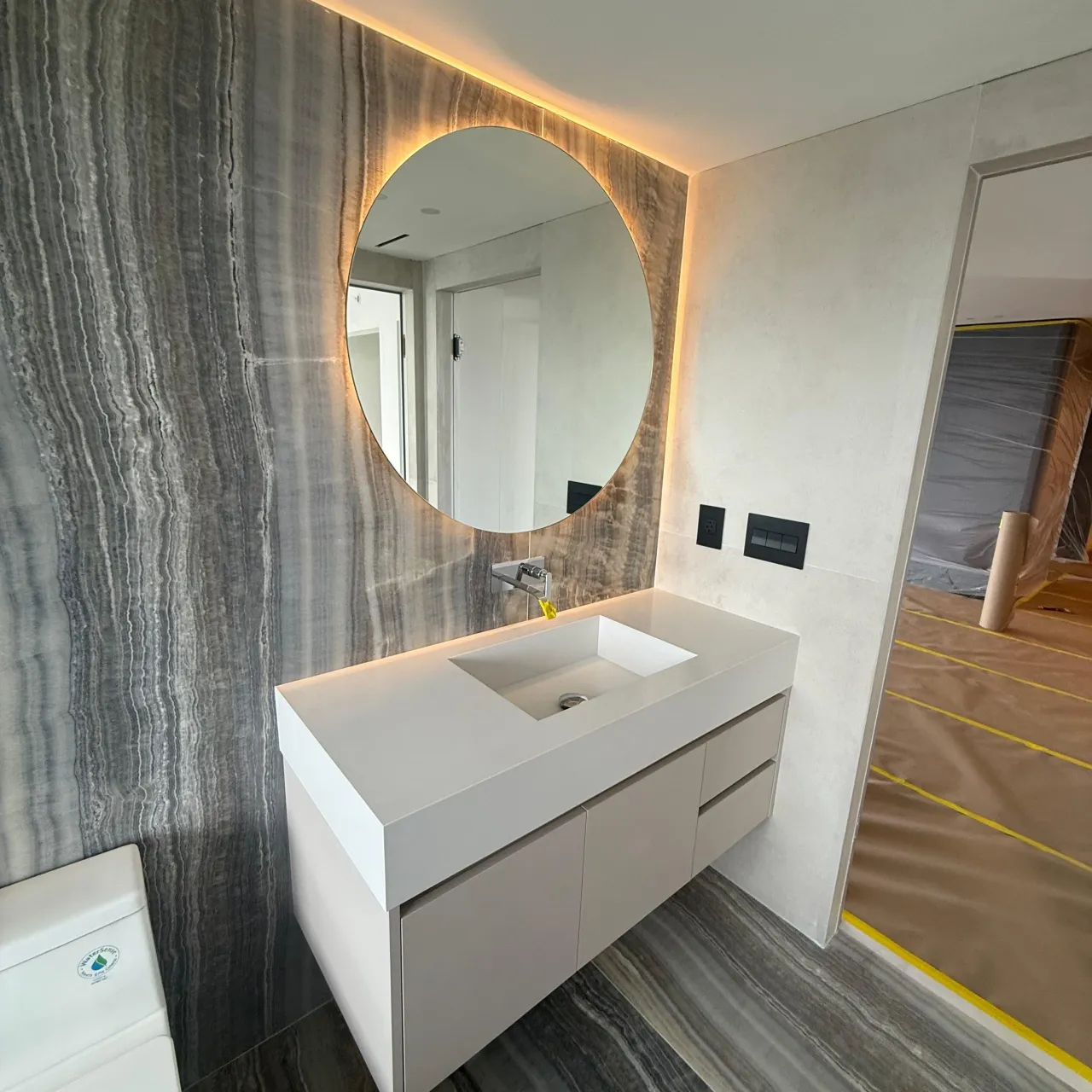 White floating vanity with an integrated sink and light countertop set against a gray-veined stone accent wall, under a round backlit mirror