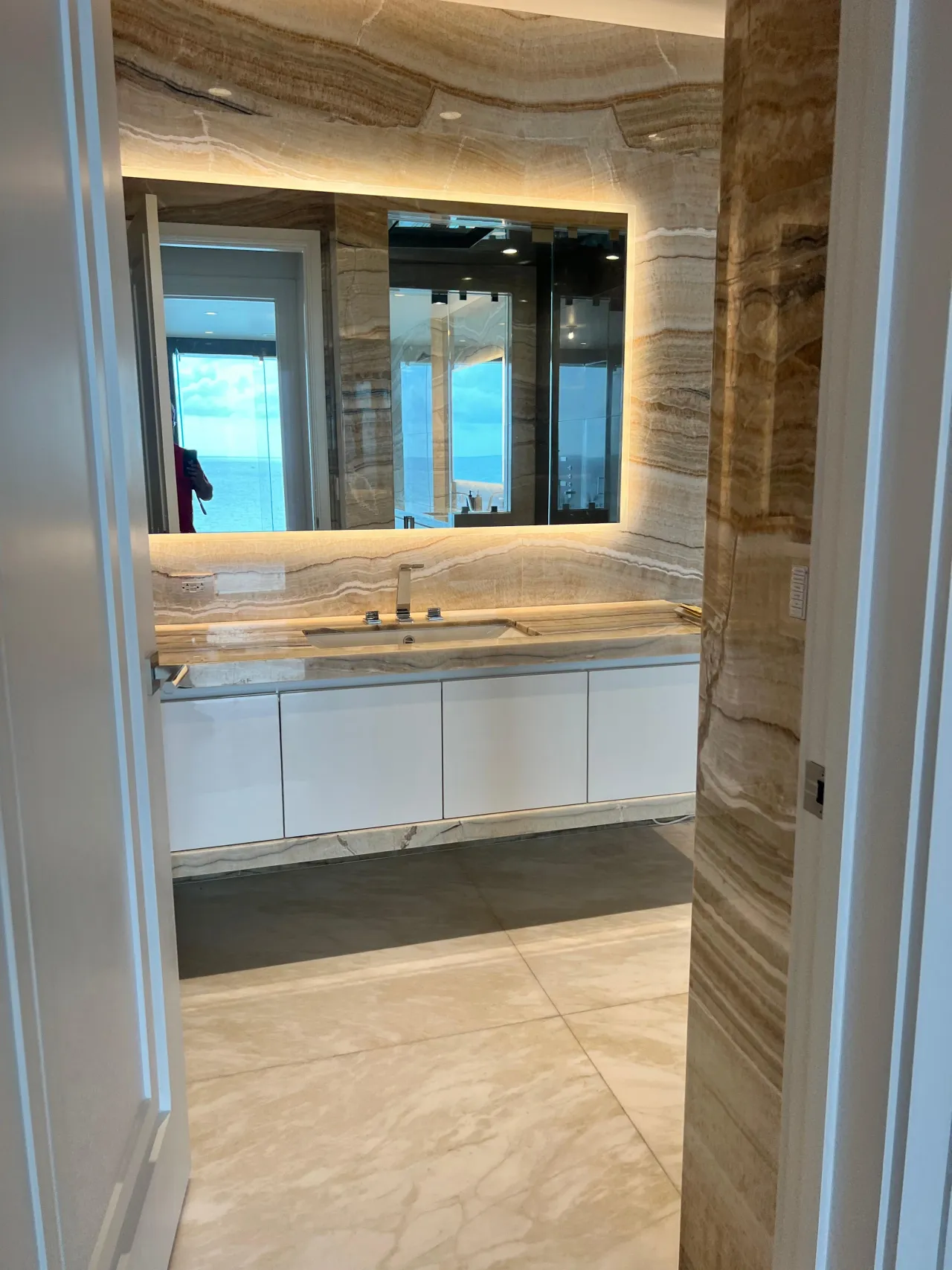 White floating vanity with a stone countertop and integrated sink beneath a large backlit mirror set into a warm brown veined-stone wall