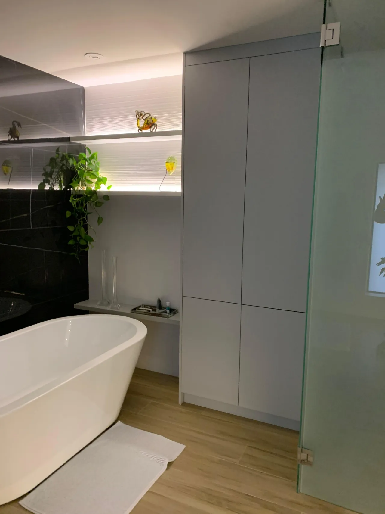 White freestanding tub beside a black-tiled accent wall with floating shelves, next to a tall light-gray storage cabinet