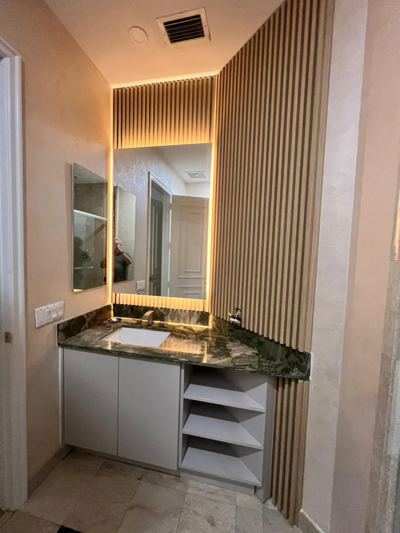 White vanity with a single undermount sink and a green-toned stone countertop, paired with open diagonal-shelf storage and a backlit mirror