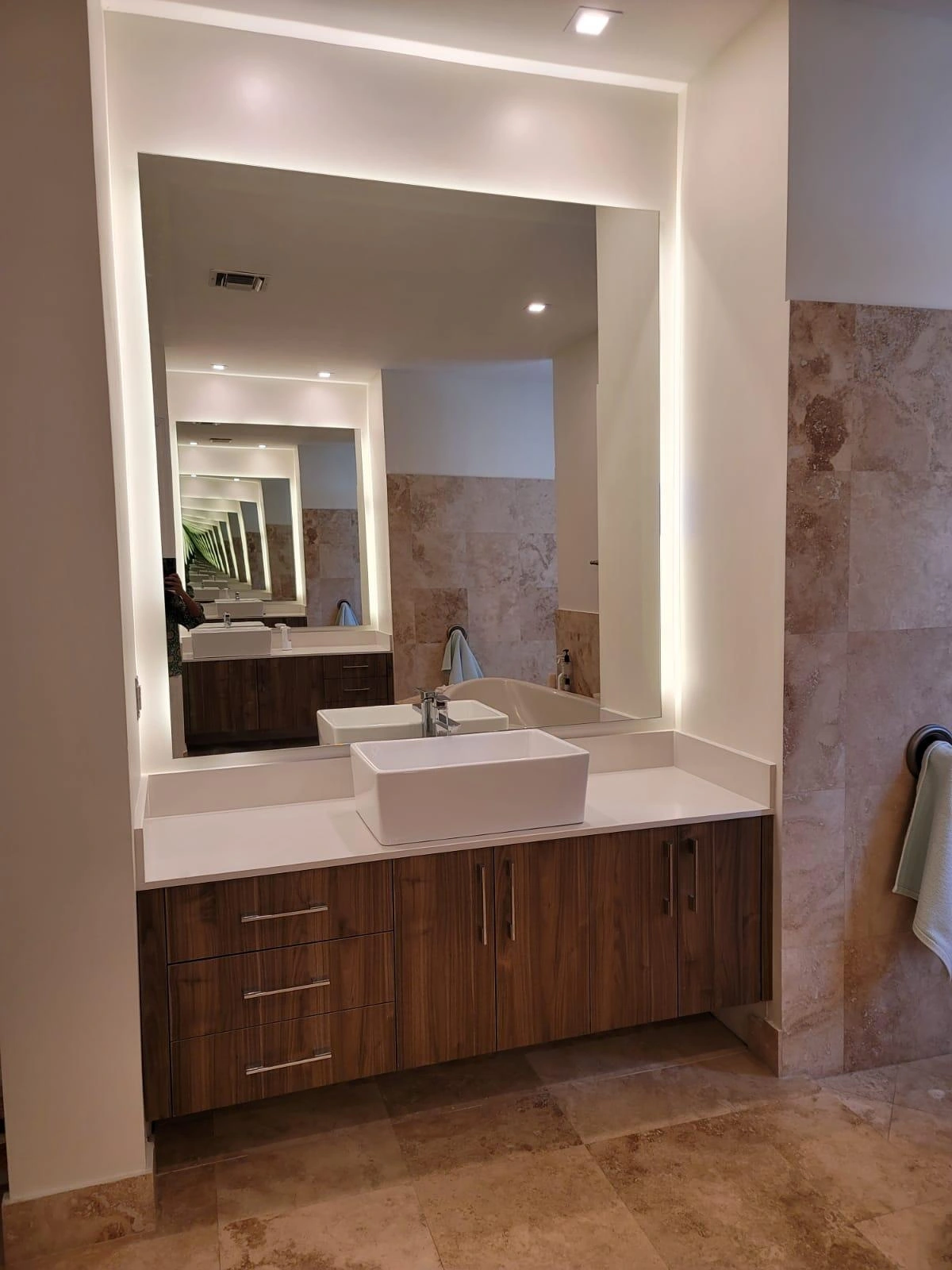 Wood-grain vanity with a white countertop, a white rectangular vessel sink and chrome faucet, below a large backlit mirror recessed in the wall