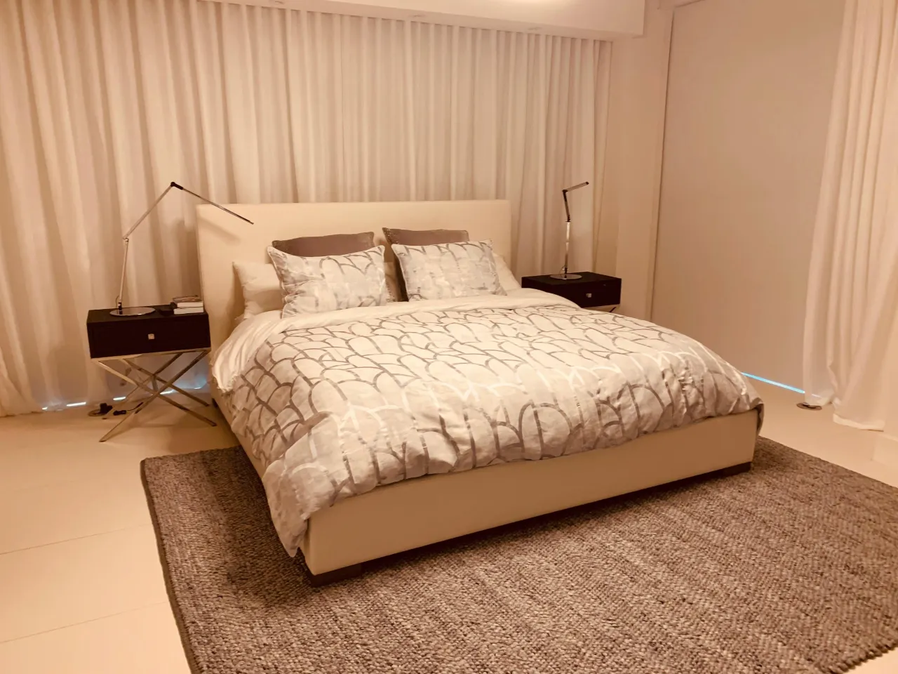 Bed with a plain cream upholstered headboard and platform base, gray and white patterned duvet, dark nightstands with reading lamps