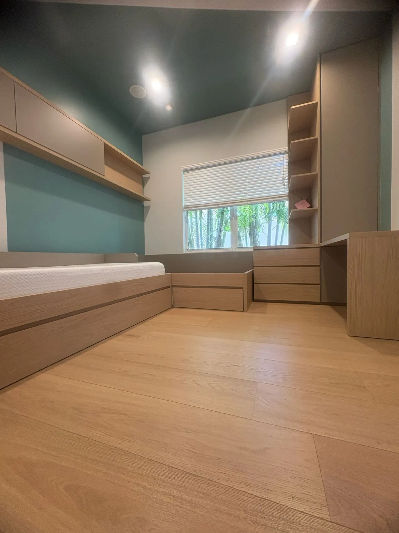 Built-in bed platform with bare mattress under floating wood-grain shelves, teal accent wall and ceiling, drawer units and tall open shelving by a window