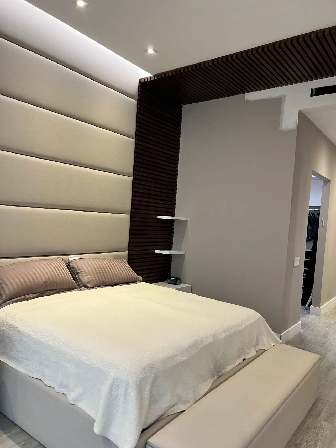 Cream channel-tufted upholstered headboard wall bordered by a vertical dark wood slat panel, with a walk-in closet visible through an open doorway
