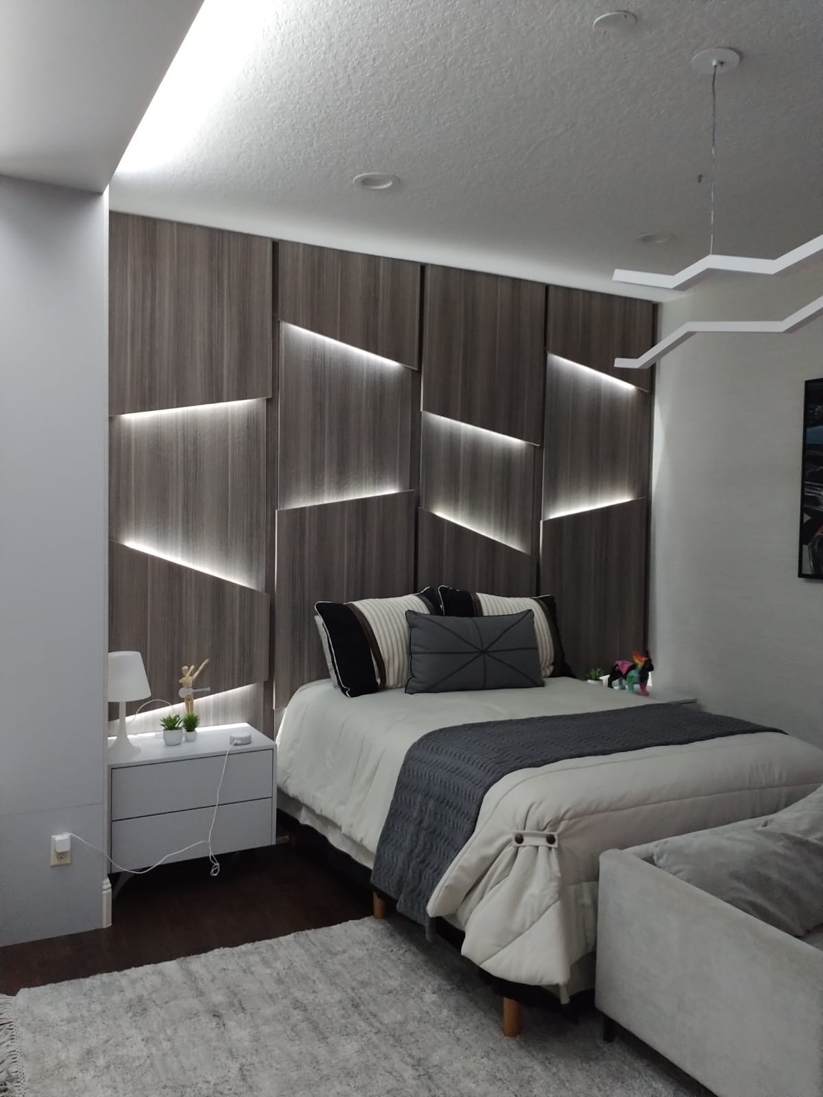 Dark wood panel wall with diagonal backlit LED cutouts behind a bed with cream/gray bedding and a nearby gray sofa