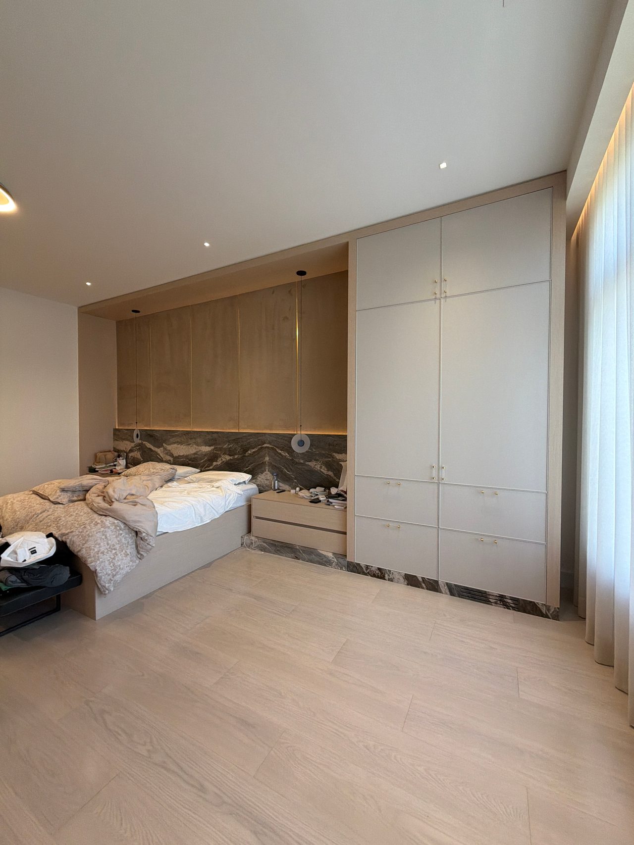 Wide view of a bedroom showing floor-to-ceiling cream closet doors, wood-grain overhead cabinets, a veined stone wall behind the bed, light wood-look floor