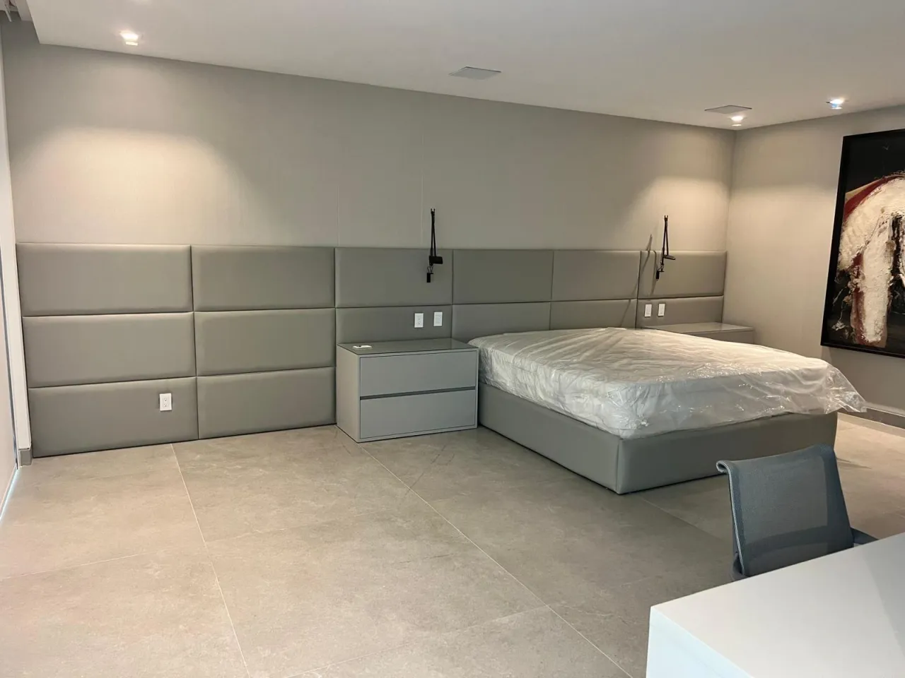 Gray padded wall panels in a grid behind a bed with plastic-wrapped mattress, matching gray two-drawer nightstand, black swing-arm sconces