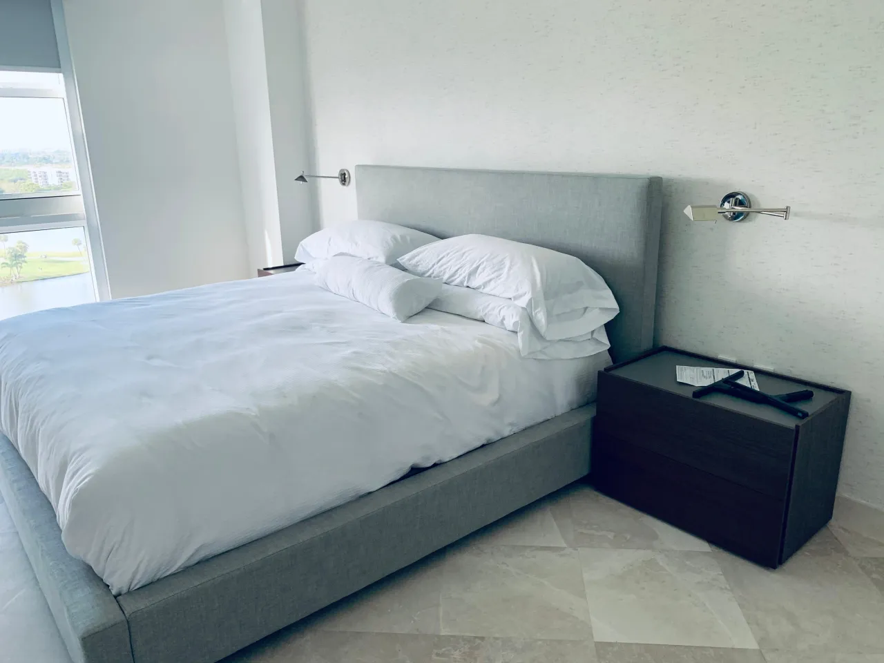 Gray upholstered platform bed with white bedding beside a dark wood nightstand, floor-to-ceiling window overlooking water
