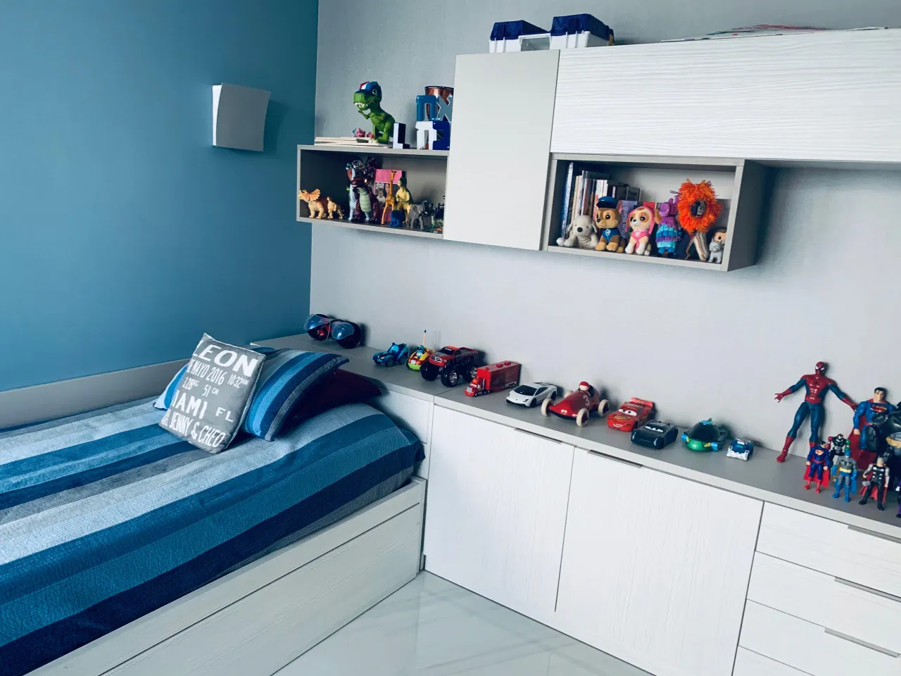 Kids bedroom with blue accent wall, white floating shelves holding toys, and white built-in cabinets below a striped bed