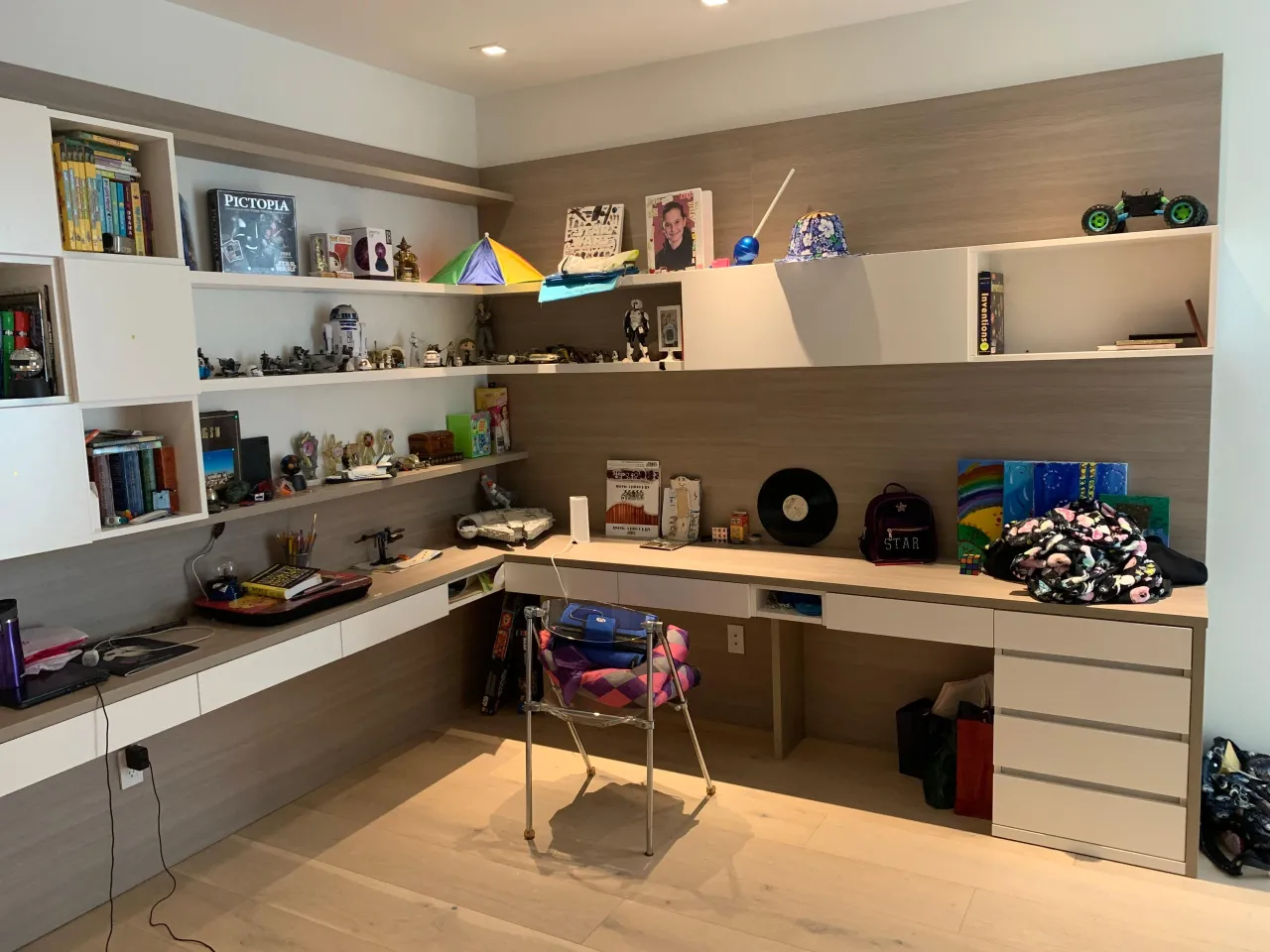 Kids L-shaped built-in desk with wood back panel beneath floating display shelves filled with books and toys