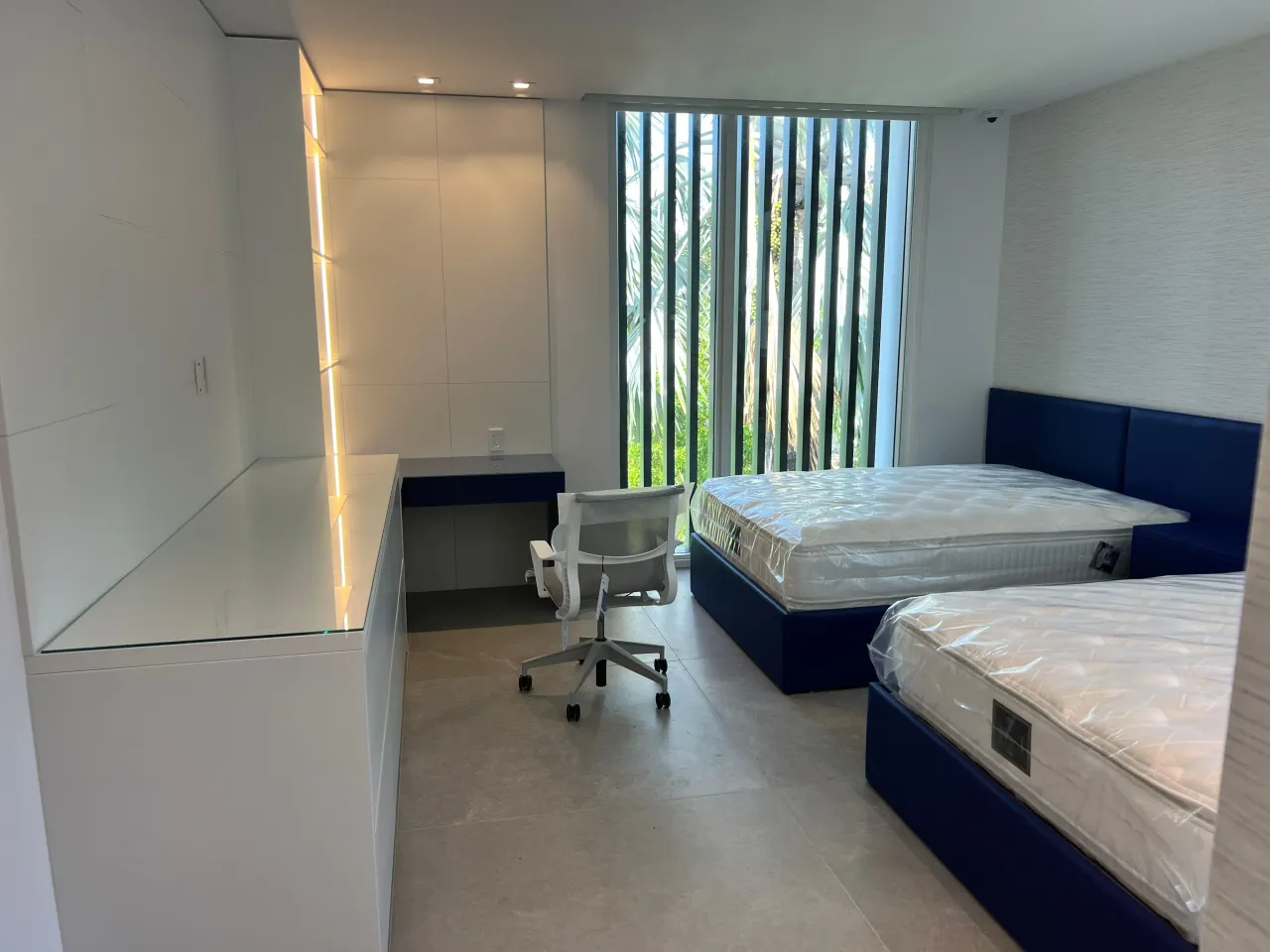Two navy upholstered beds with plastic-wrapped mattresses, white built-in desk and LED-lit shelving, floor-to-ceiling window with vertical blinds