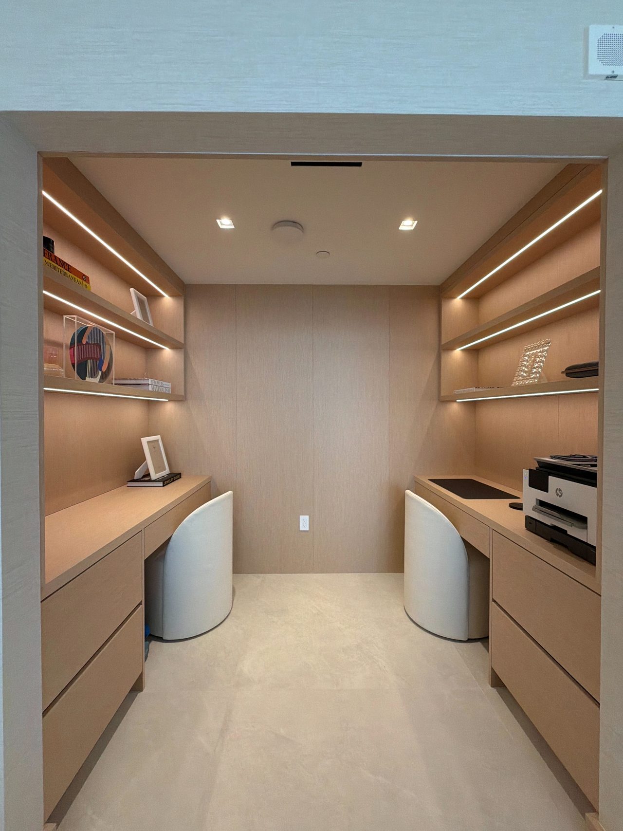Office nook with two facing veneer desks, lit open shelving above each, drawer banks, and curved white chairs under recessed lights