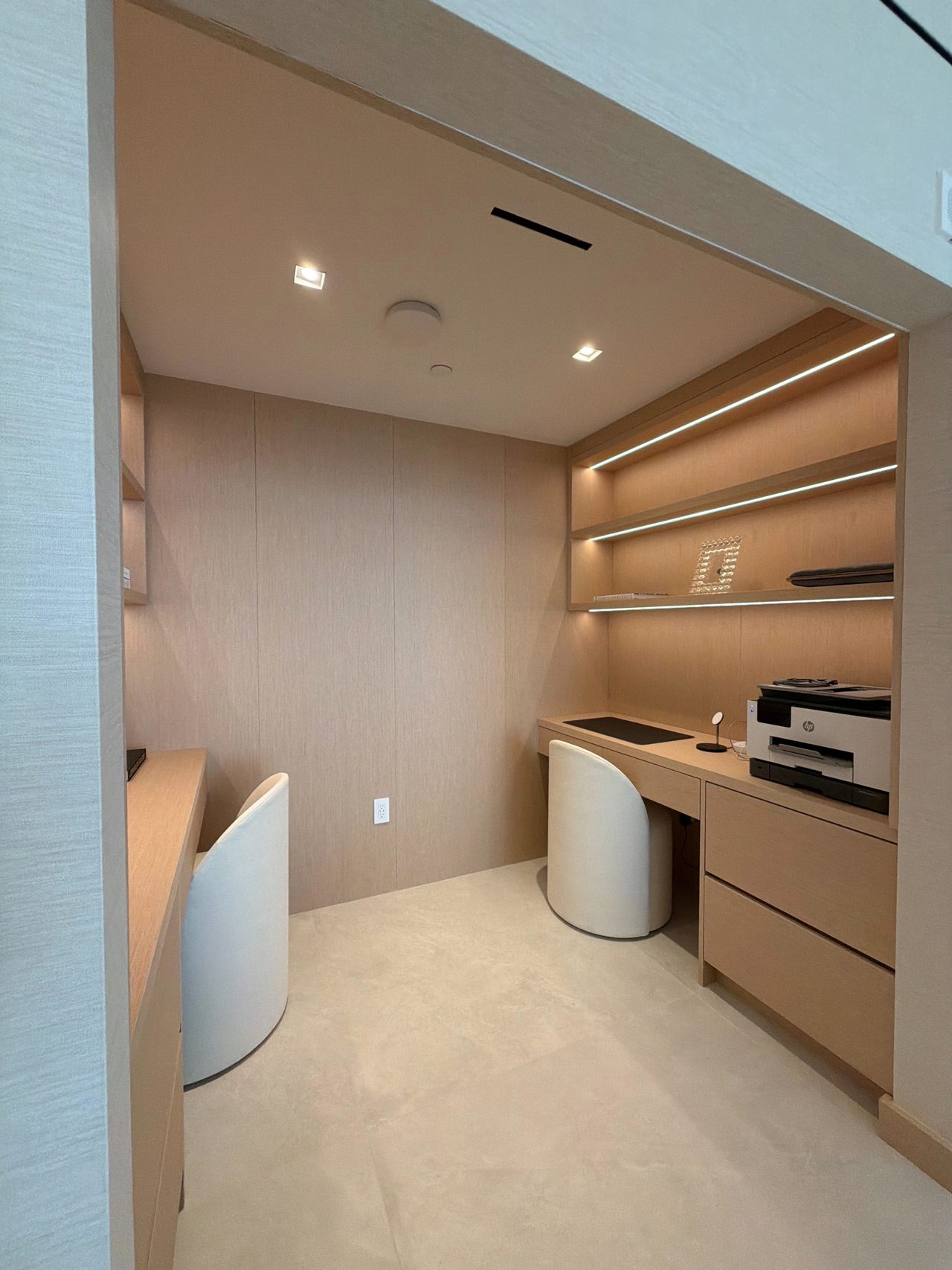 Veneer office nook desk with a drawer bank under stacked integrated-LED shelves, a curved white chair, and recessed ceiling lights