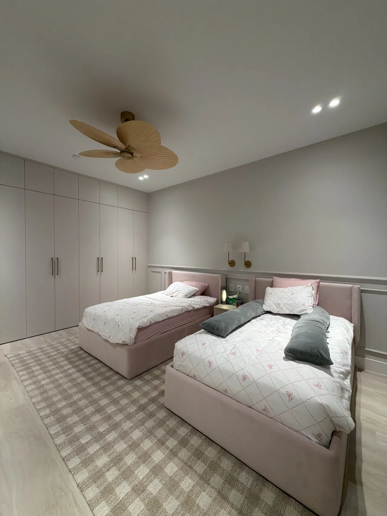 Pink upholstered twin beds on a checked gingham rug, gold-tone sconces above the headboards, woven palm-blade ceiling fan, tall closet doors